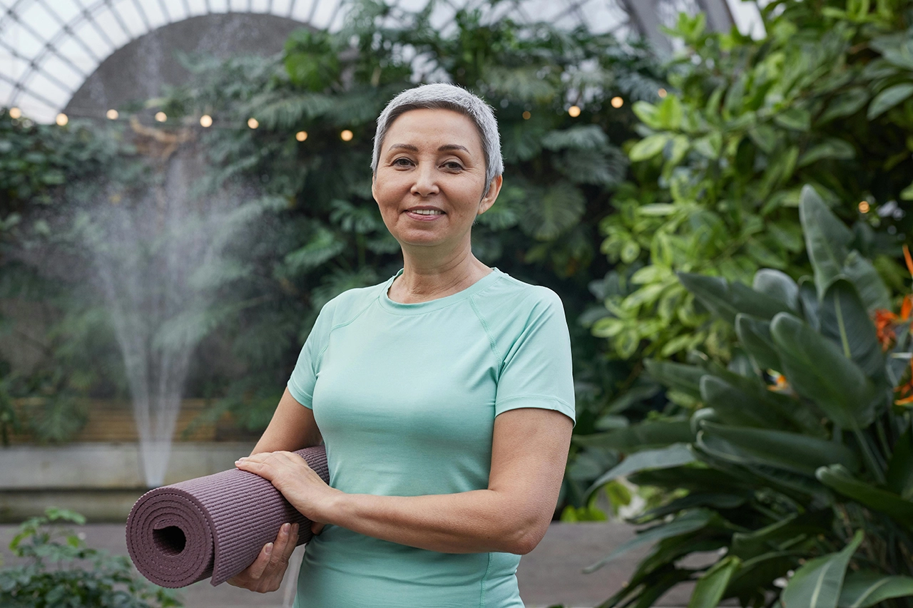 Healthy, mature women holding a yoga mat with trees and landscaping behind her.