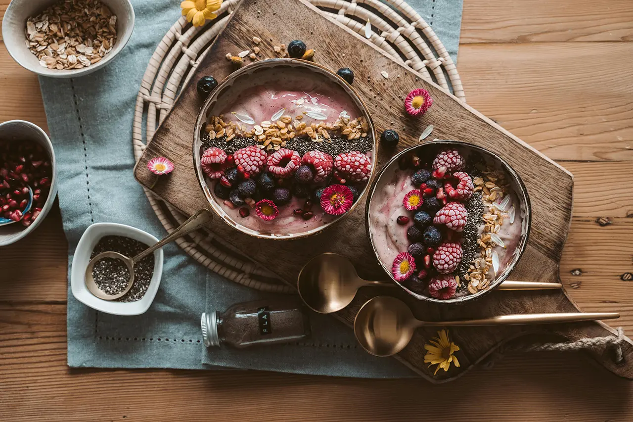 top view of two bowls of acai with chia seeds, berries and granola sitting on a cutting board with flowers and ingredients in bowls surrounding it.