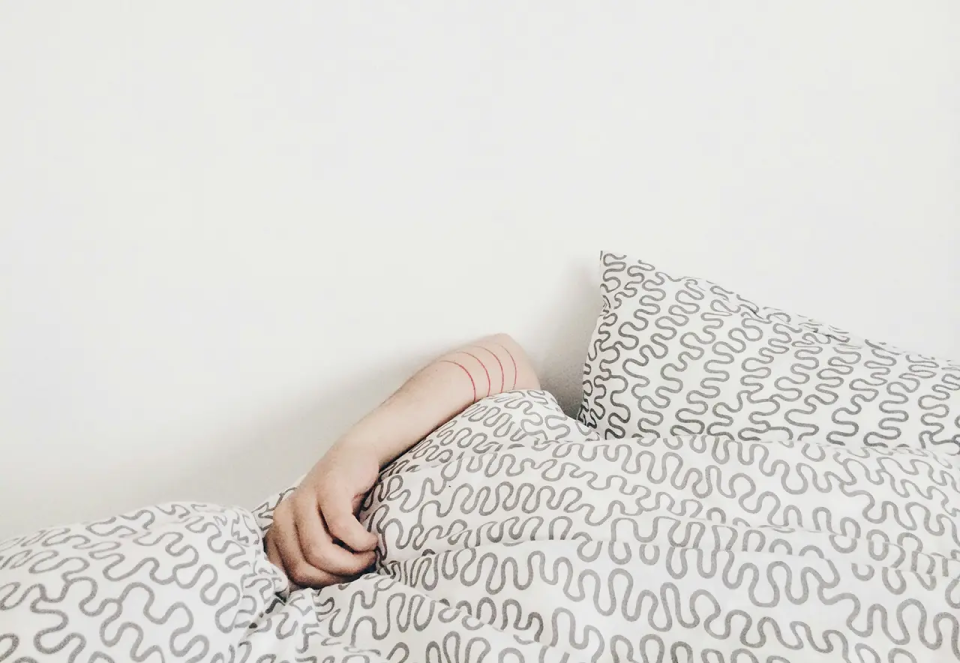 A person’s arm sticking out from under a white blanket with gray squiggly patterns, lying in bed against a plain white wall.